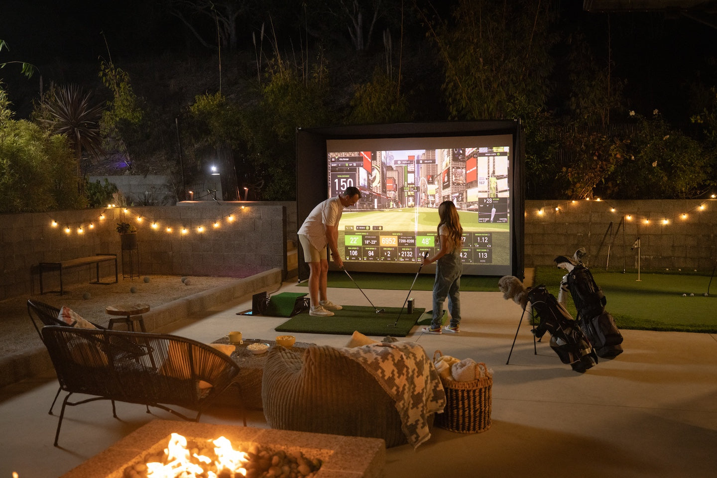 Outdoor backyard golf simulator setup at night with SKYTRAK launch monitor, large projection screen, string lights, fire pit, and patio seating
