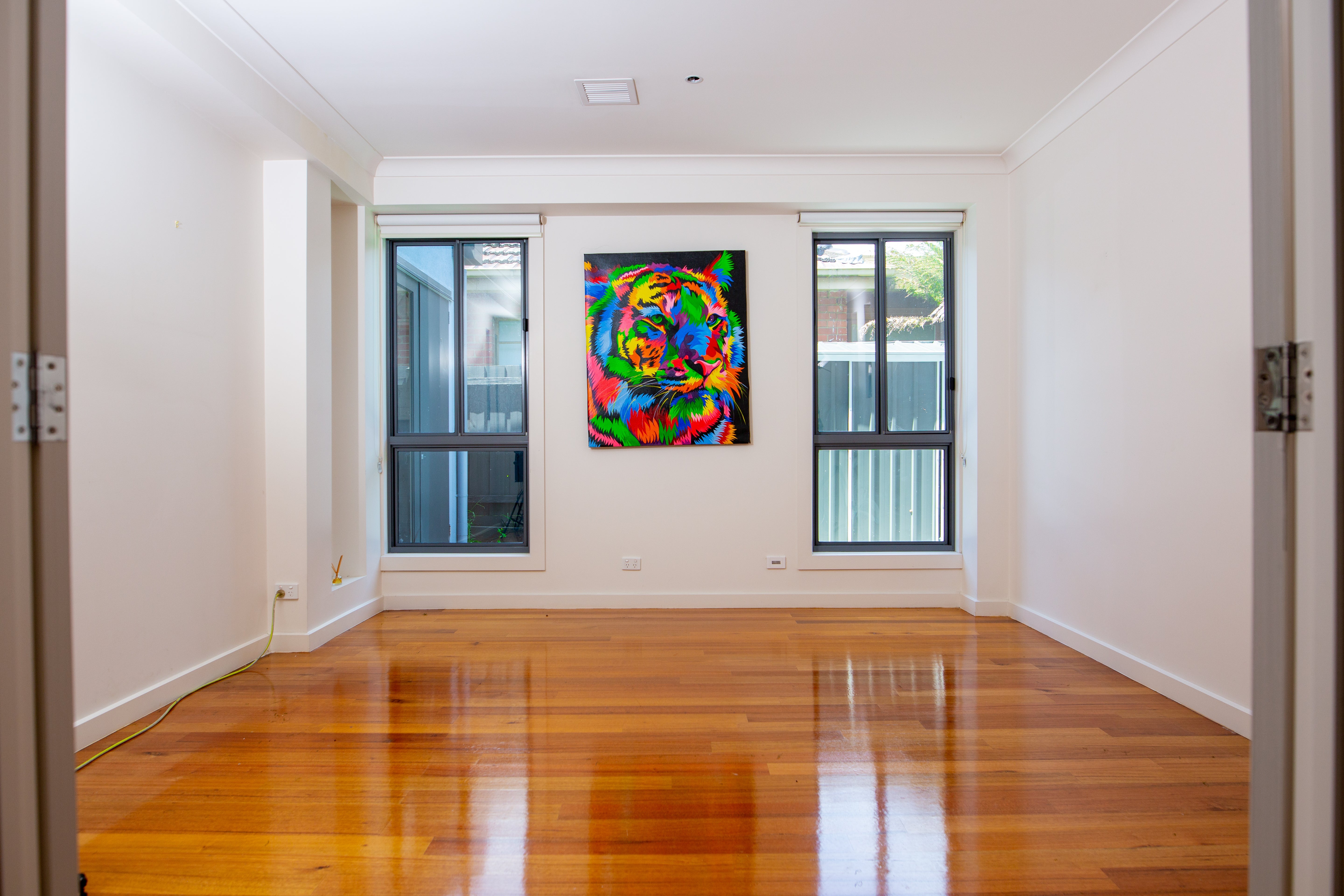 Bright, modern minimalist interior room featuring a vibrant, colorful abstract tiger painting as a focal point on a clean white wall, positioned between two large windows above natural wood flooring. Perfect setup for a full home simulator setup!